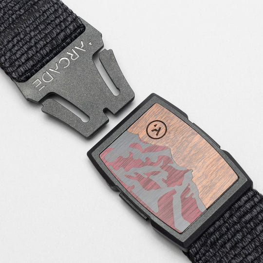 A close-up of the Arcade Belts Woody buckle, featuring a mountain landscape and smiley face design, attached to a woven black elastic stretch belt on a wood-like background.
