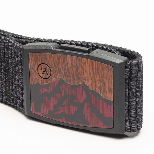A close-up of the Woody belt by Arcade Belts, featuring black woven elastic, a rectangular buckle with a woodgrain and mountain silhouette in dark red and black, plus a small circular logo in the corner.
