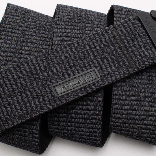 A close-up photo of the Arcade Belts Woody, a black woven fabric belt featuring a rectangular ARCADE logo patch. The belt’s stretch webbing pattern and texture are prominently displayed.