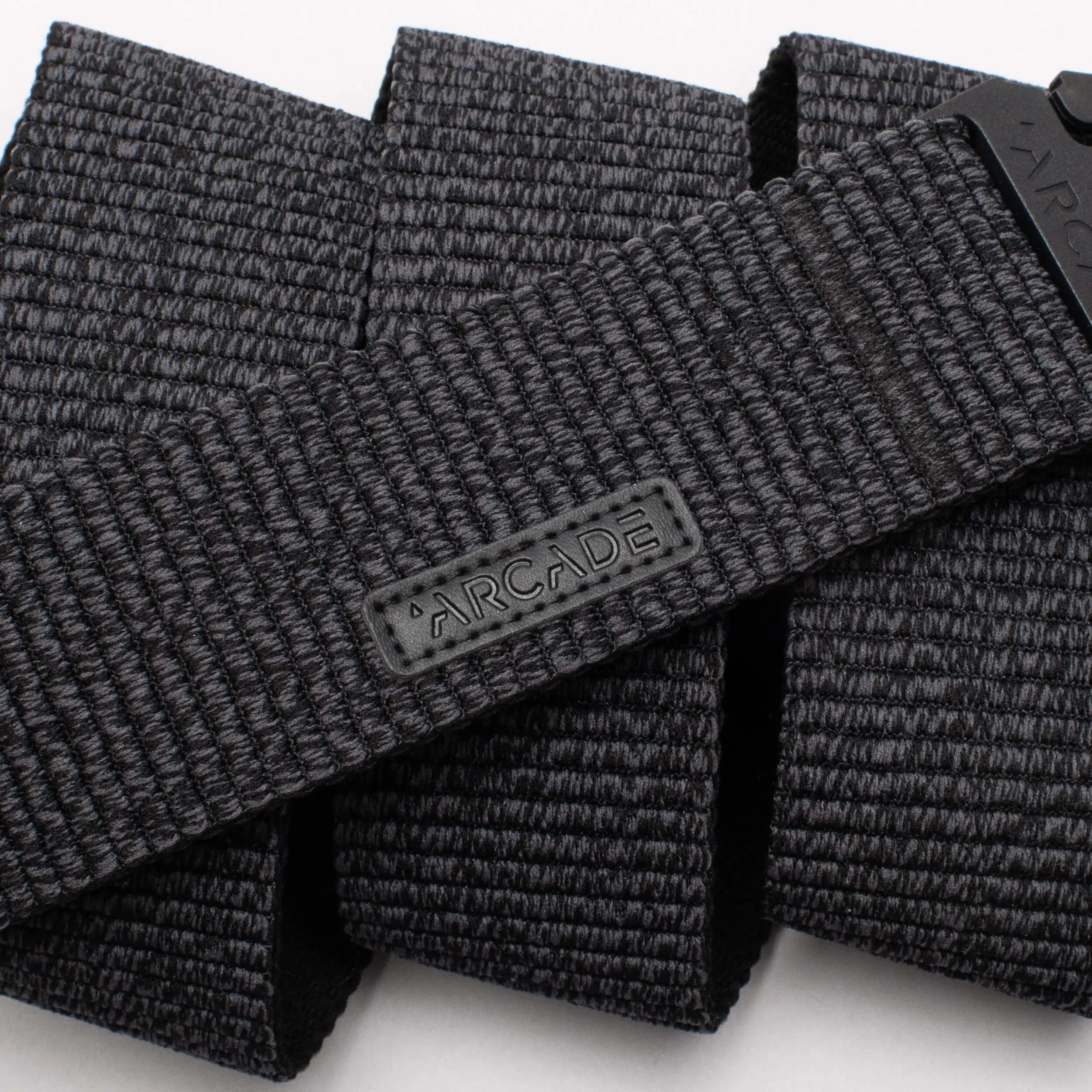 A close-up photo of the Arcade Belts Woody, a black woven fabric belt featuring a rectangular ARCADE logo patch. The belt’s stretch webbing pattern and texture are prominently displayed.