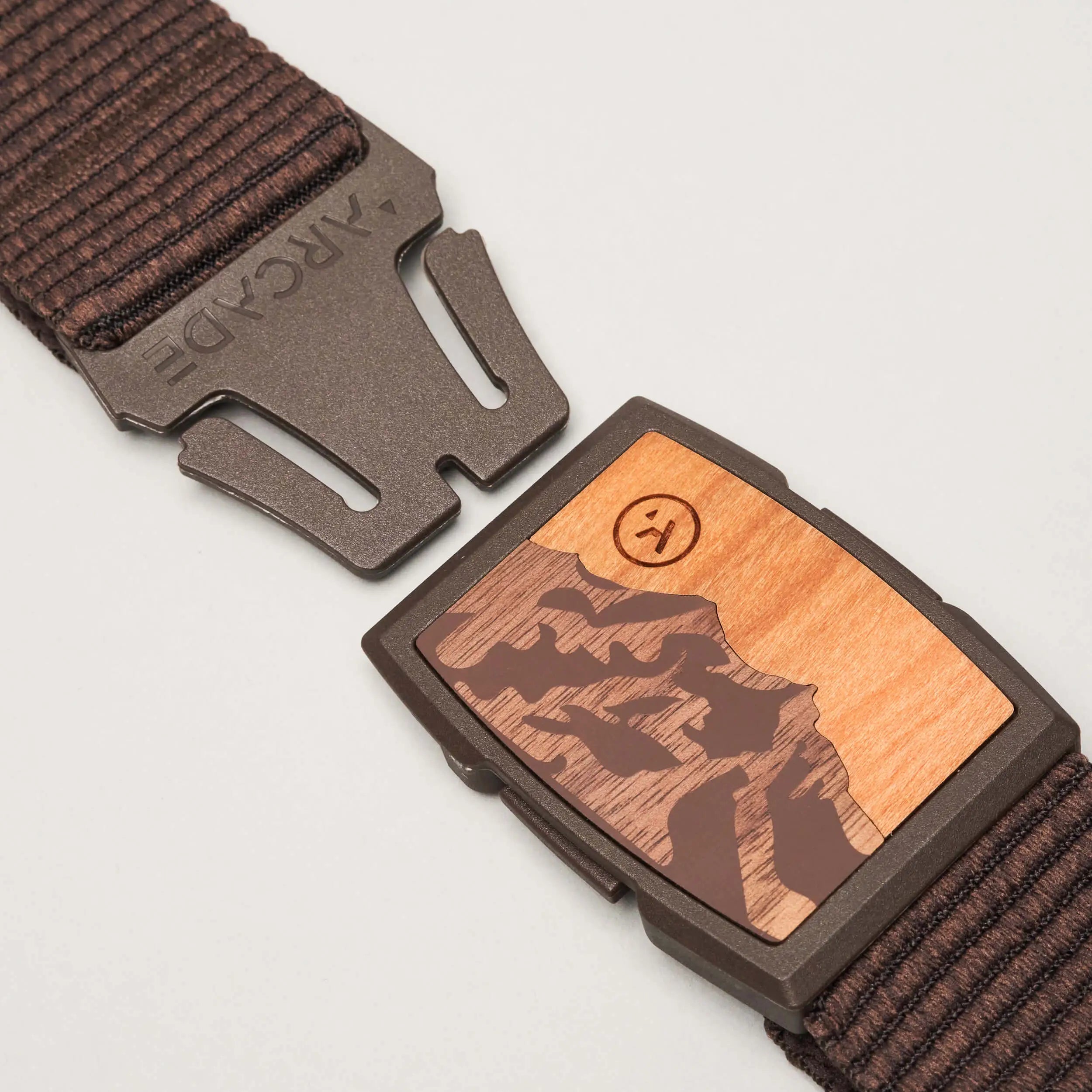 A close-up of the Arcade Belts Woody shows its brown woven design and partially open metal clasp with a wooden inlay engraved with a mountain scene and a smiley face. The low-profile Woody belt seamlessly blends style and function.