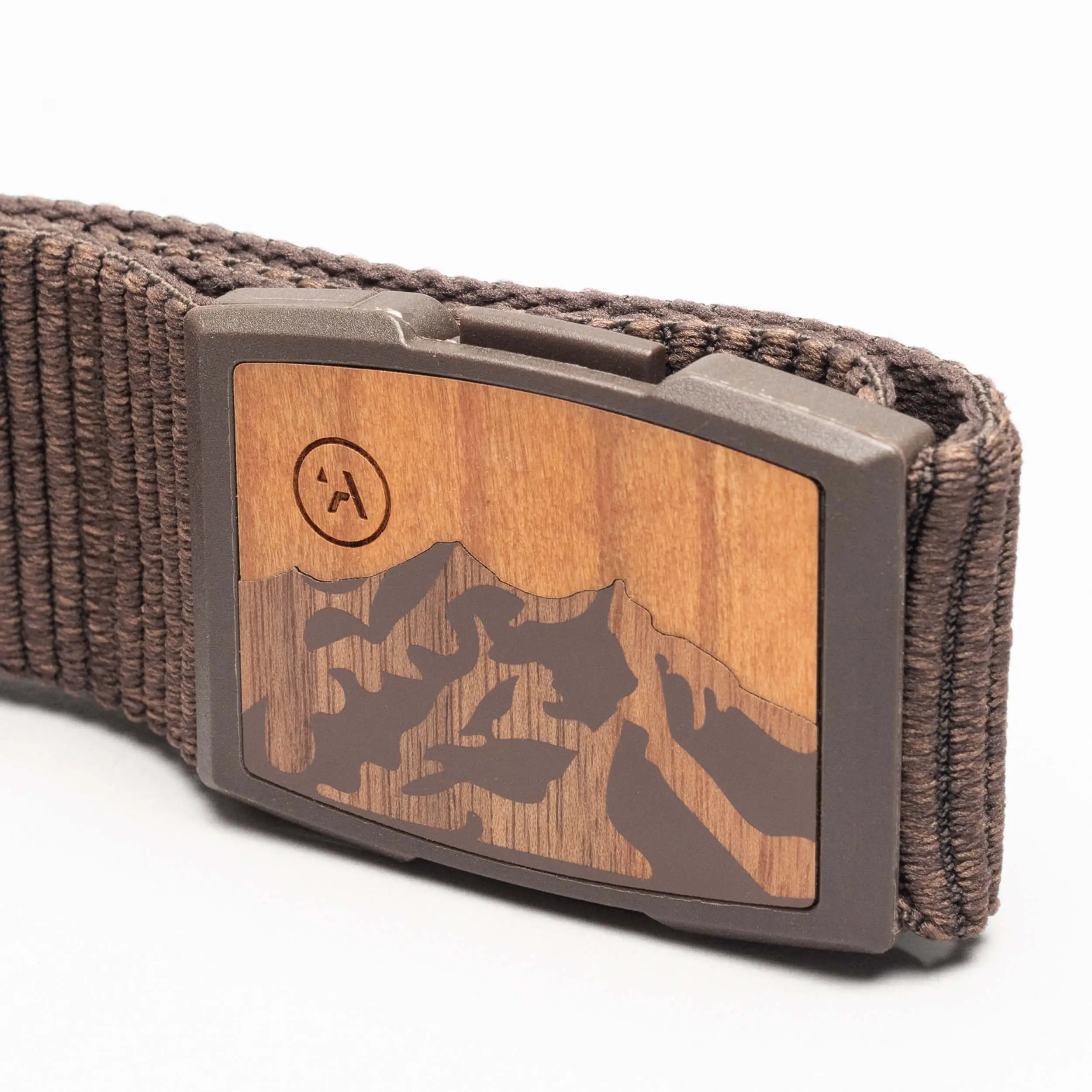 A close-up of the Arcade Belts Woody: a brown elastic stretch belt with a woven design and a rectangular wood-finish buckle engraved with a mountain motif. The brand’s encircled “A” logo appears on the upper left of the buckle.