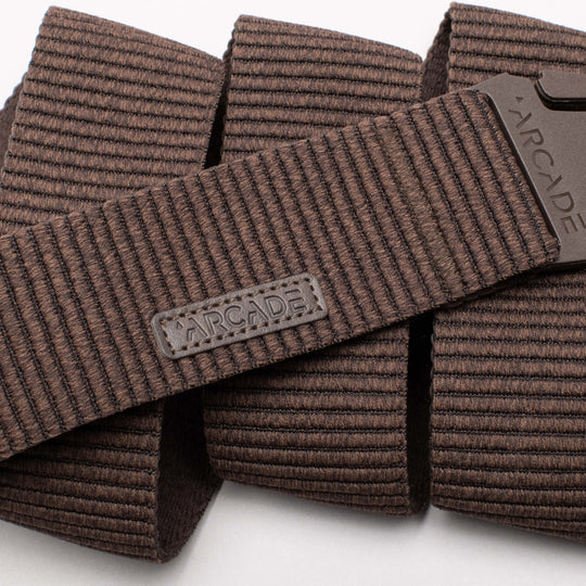 Close-up of the Arcade Belts Woody, a brown textured elastic stretch belt with a rectangular ARCADE logo patch and matching brown plastic buckle, both displaying the brand name. The coiled belt highlights its flexible stretch webbing.