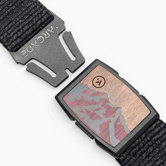 A close-up of the Arcade Belts Woody buckle, featuring a mountain landscape and smiley face design, attached to a woven black elastic stretch belt on a wood-like background.