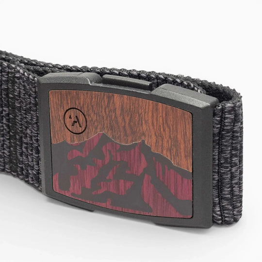 A close-up of the Woody belt by Arcade Belts, featuring black woven elastic, a rectangular buckle with a woodgrain and mountain silhouette in dark red and black, plus a small circular logo in the corner.