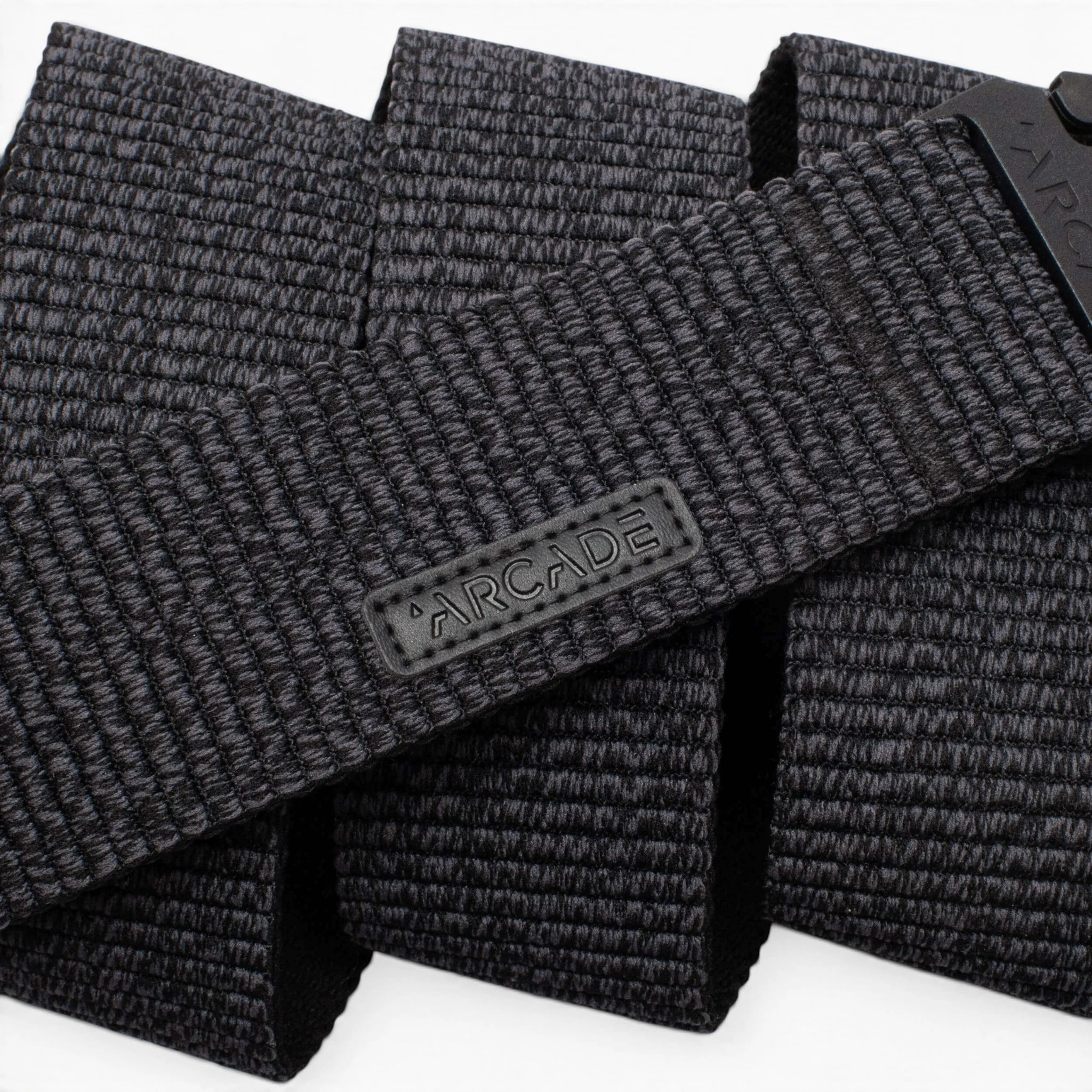 A close-up photo of the Arcade Belts Woody, a black woven fabric belt featuring a rectangular ARCADE logo patch. The belt’s stretch webbing pattern and texture are prominently displayed.