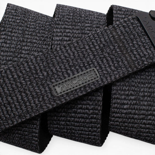 A close-up photo of the Arcade Belts Woody, a black woven fabric belt featuring a rectangular ARCADE logo patch. The belt’s stretch webbing pattern and texture are prominently displayed.