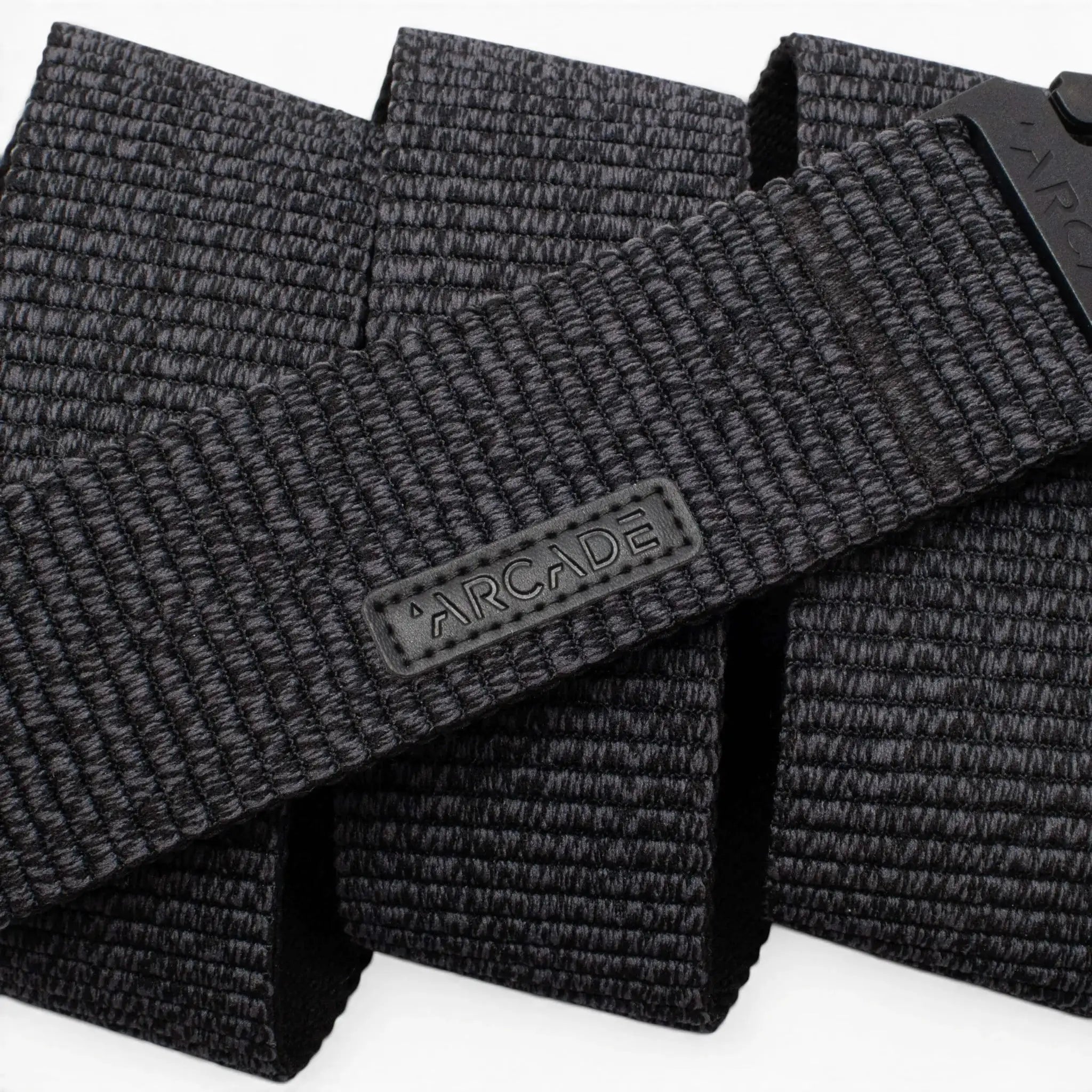 A close-up photo of the Arcade Belts Woody, a black woven fabric belt featuring a rectangular ARCADE logo patch. The belt’s stretch webbing pattern and texture are prominently displayed.