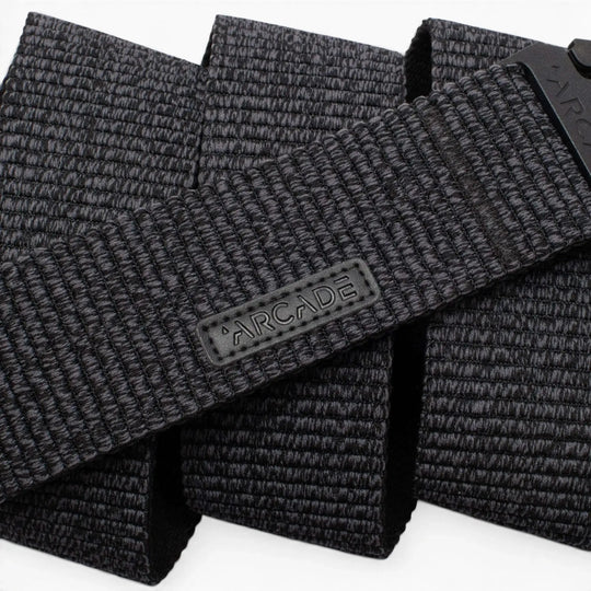 A close-up photo of the Arcade Belts Woody, a black woven fabric belt featuring a rectangular ARCADE logo patch. The belt’s stretch webbing pattern and texture are prominently displayed.