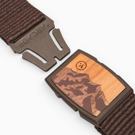 A close-up of the Arcade Belts Woody shows its brown woven design and partially open metal clasp with a wooden inlay engraved with a mountain scene and a smiley face. The low-profile Woody belt seamlessly blends style and function.