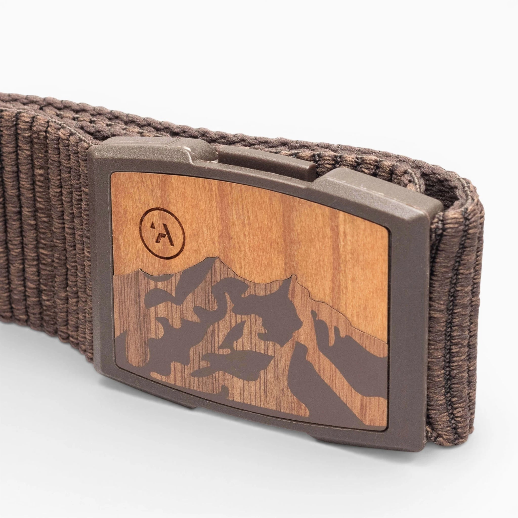 A close-up of the Arcade Belts Woody: a brown elastic stretch belt with a woven design and a rectangular wood-finish buckle engraved with a mountain motif. The brand’s encircled “A” logo appears on the upper left of the buckle.