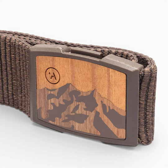 A close-up of the Arcade Belts Woody: a brown elastic stretch belt with a woven design and a rectangular wood-finish buckle engraved with a mountain motif. The brand’s encircled “A” logo appears on the upper left of the buckle.