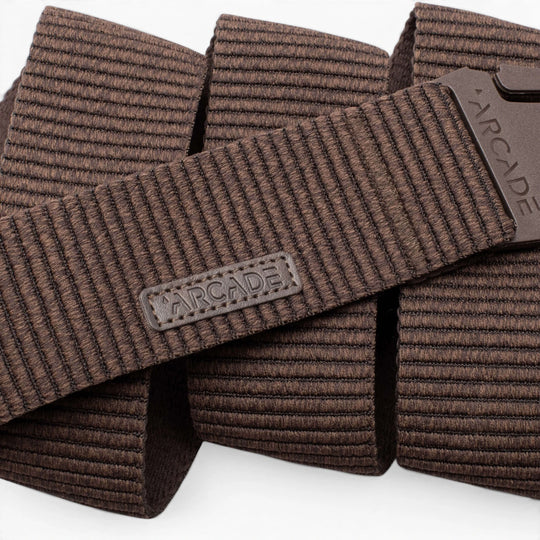 Close-up of the Arcade Belts Woody, a brown textured elastic stretch belt with a rectangular ARCADE logo patch and matching brown plastic buckle, both displaying the brand name. The coiled belt highlights its flexible stretch webbing.