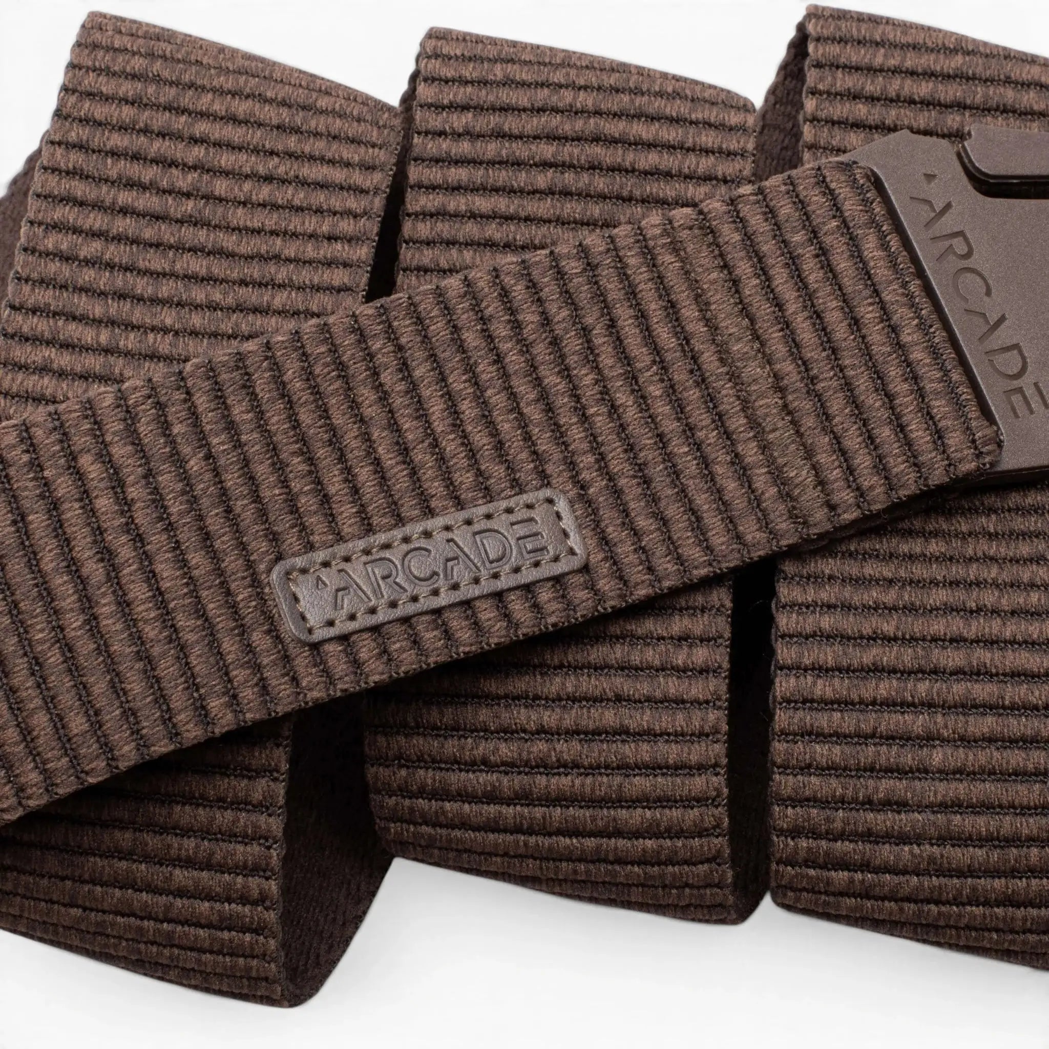 Close-up of the Arcade Belts Woody, a brown textured elastic stretch belt with a rectangular ARCADE logo patch and matching brown plastic buckle, both displaying the brand name. The coiled belt highlights its flexible stretch webbing.