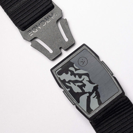 The Woody by Arcade Belts is a black elastic stretch belt with a matte black buckle and a rectangular metal plate featuring an abstract mountain design plus a small circular logo on the right.