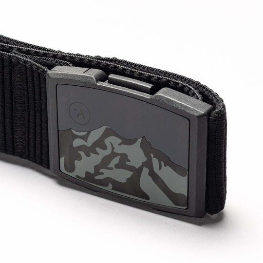 Close-up of the Arcade Belts Woody elastic stretch belt with a metal buckle featuring a mountain range design and an etched moon or sun in the corner. The white background highlights this low-profile belt.