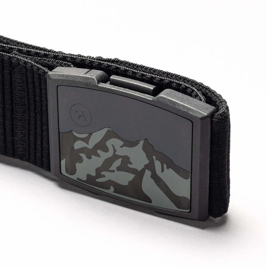 Close-up of the Arcade Belts Woody elastic stretch belt with a metal buckle featuring a mountain range design and an etched moon or sun in the corner. The white background highlights this low-profile belt.