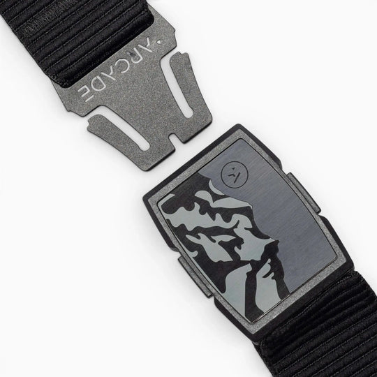 The Woody by Arcade Belts is a black elastic stretch belt with a matte black buckle and a rectangular metal plate featuring an abstract mountain design plus a small circular logo on the right.