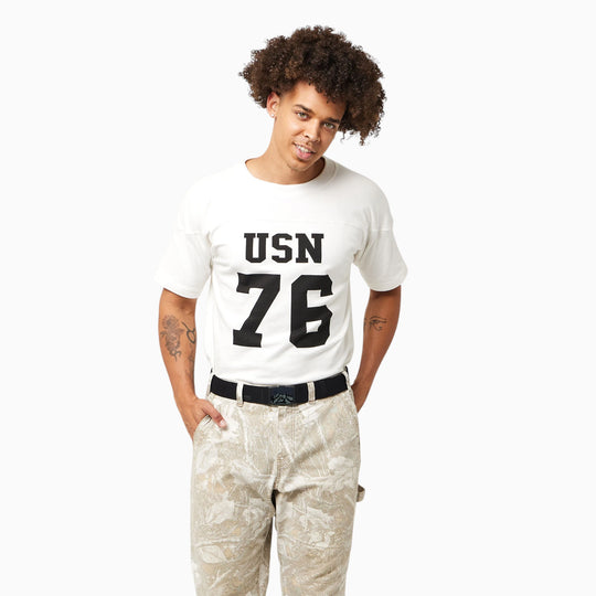 A person with curly hair stands against a plain white background wearing a white USN 76 t-shirt, patterned beige pants, and an Arcade Belts Woody Kit belt featuring a wood inlay buckle in Woody Heather Walnut.