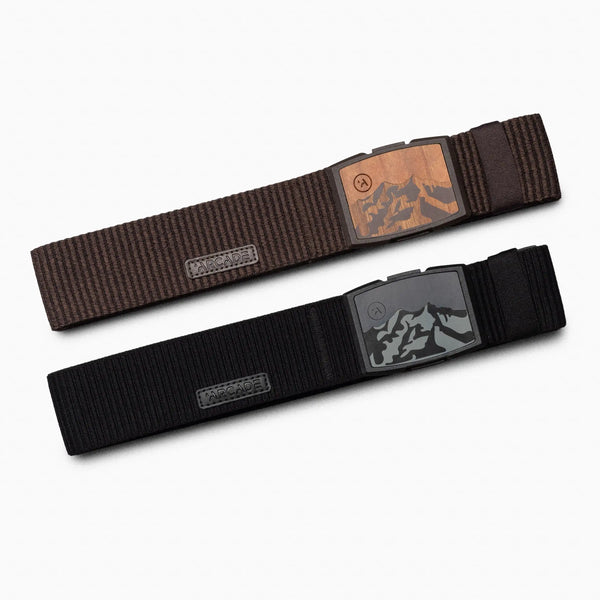 The Arcade Belts Woody Kit features two fabric belts laid flat on a white background: one dark brown with a wood-look plastic buckle, the other black with a mountain design. Both belts are made from elastic stretch webbing for comfort.