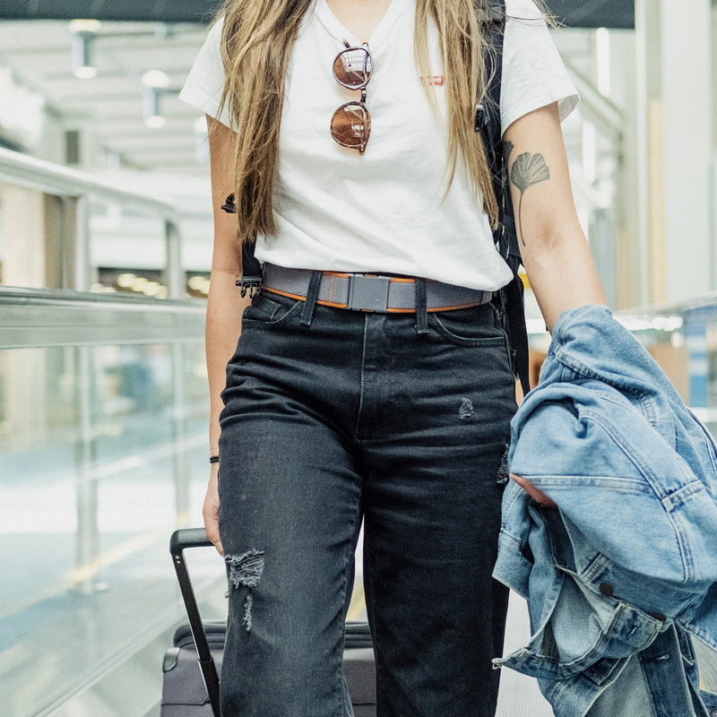 A person wearing a white t-shirt, black ripped jeans, and sunglasses on their shirt walks through an airport, holding a denim jacket and pulling a suitcase. Only the torso and legs are visible.