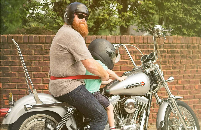 A bearded man wearing sunglasses and a helmet, secured with a durable Arcade Belt and plastic buckle, rides a silver motorcycle with a young child in front. They are next to a brick wall with trees in the background.