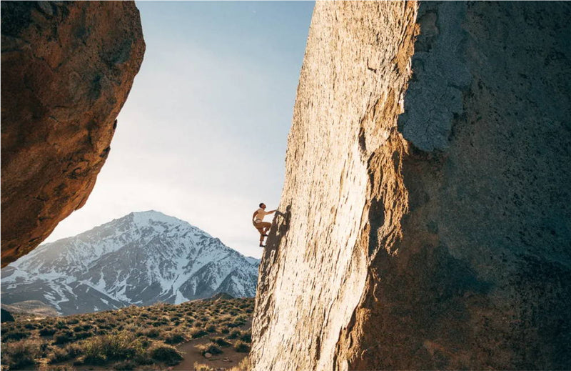 A person rock climbing a steep, large boulder with Stretch Webbing gear, against a snowy mountain and clear sky. Sunlight casts shadows, highlighting the rugged terrain and dramatic landscape.