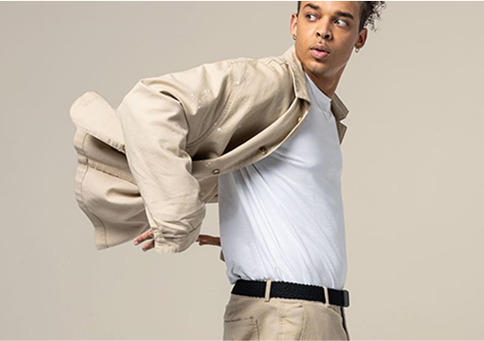 A person wearing a white t-shirt and beige pants is slipping on a matching beige button-up shirt, featuring an Arcade Belt with stretch webbing, looking to the side against a plain, light background.
