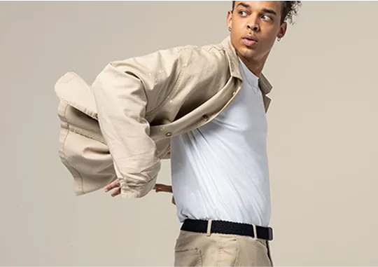 A young man wears a light beige overshirt, white t-shirt, and beige pants with a black belt. He is mid-motion, looking to his left, against a plain light background.