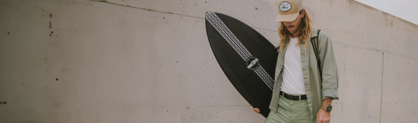 A person with long hair, wearing an olive jacket, pants secured by a Low Profile Belt, and a beige cap, walks along a concrete wall while carrying a black surfboard.