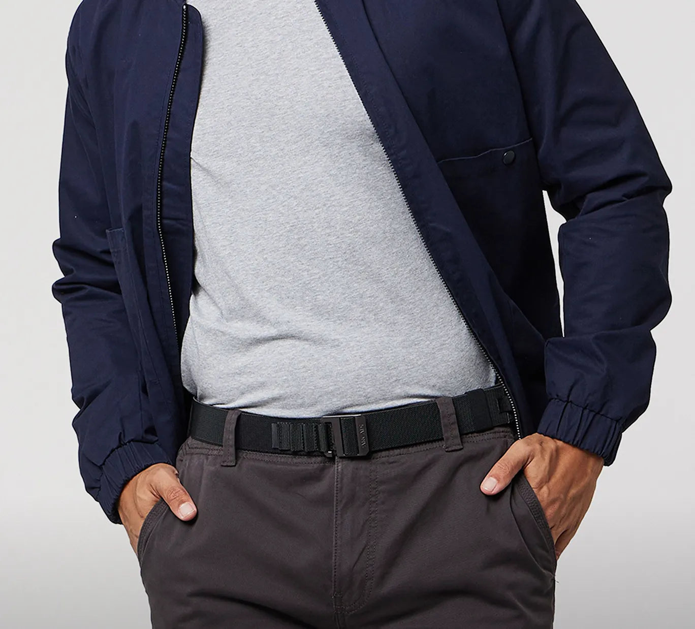 A person wearing a dark blue jacket over a light gray t-shirt, with their hands in the pockets of dark gray pants, and a black belt visible around the waist.