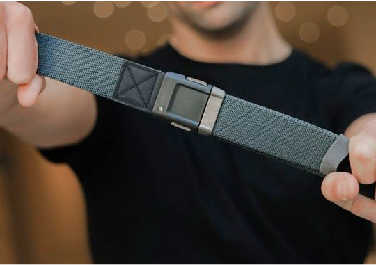 A person wearing a black shirt holds a gray stretch webbing travel belt with a rectangular metal buckle, stretching it towards the camera. The background is blurred with warm lighting.