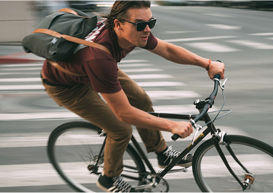 A man wearing sunglasses, a maroon shirt, and brown pants rides a bicycle quickly through a city intersection, his gray messenger bag slung over his shoulder and a low profile belt with stretch webbing for comfort around his waist.