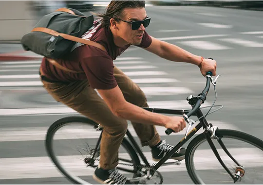 A man wearing sunglasses, a maroon shirt, and brown pants rides a bicycle quickly through a city street, carrying a messenger bag over his shoulder.
